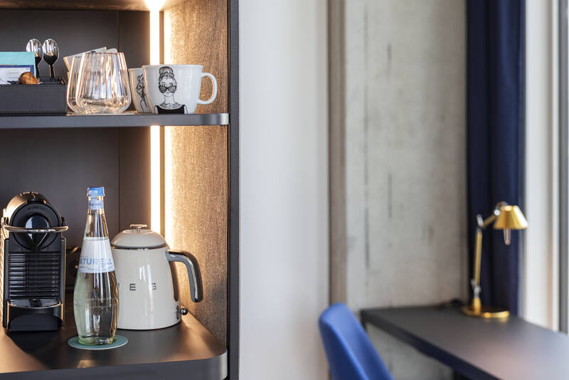beleuchteter Kaffeezubereitungsbereich mit Nespresso-Maschine, Glasflasche, weißem Wasserkocher und Tasse mit Gesichtsmotiv auf schwarzem Regal neben Schreibtisch mit blauem Stuhl und goldener Lampe