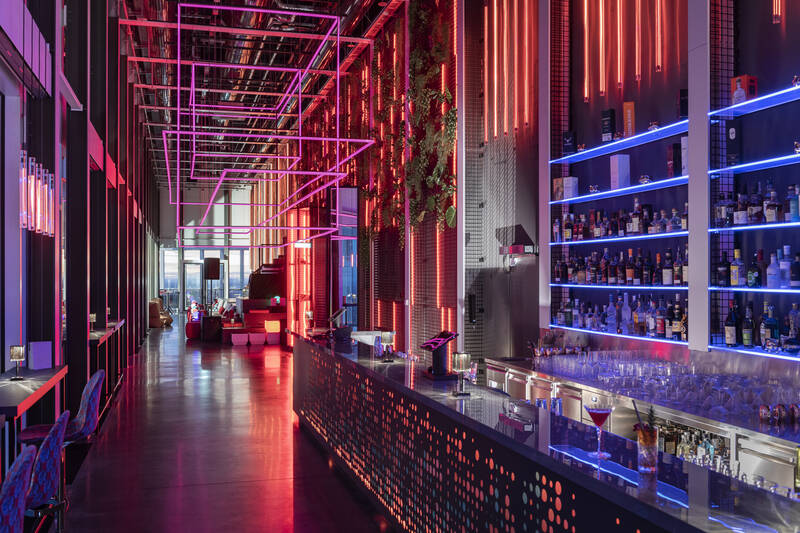 Moderne Bar mit langen Tresen, beleuchtet von pinken und blauen Neonlichtern, mit Regalen voller Flaschen und Sitzgelegenheiten im Hintergrund.