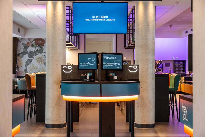 Digitaler Check-in Bereich mit Welcome Desk mit mehreren Bildschirmen und Leuchtschildern, die Begrüßung und KI-Optimierung anzeigen