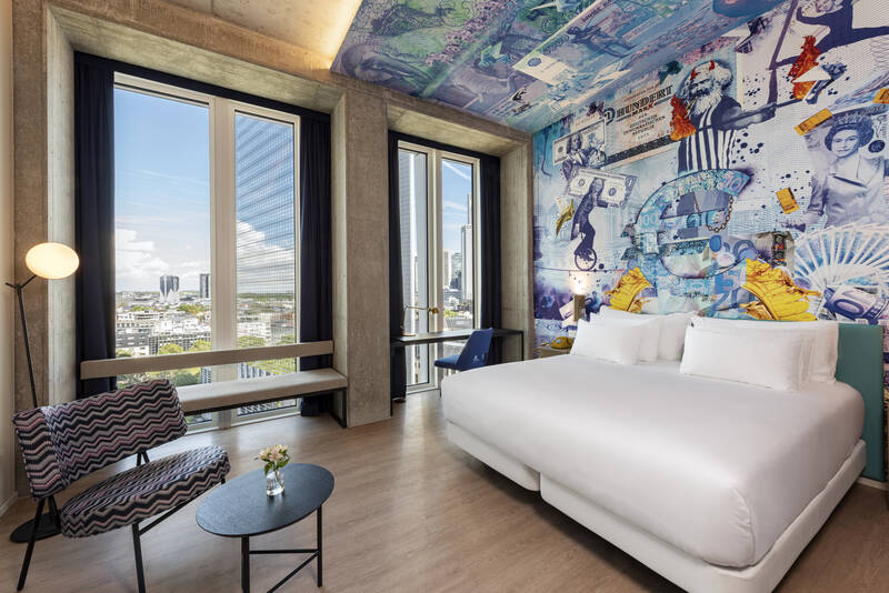 Modernes Hotelzimmer mit großem Bett, bunter Wandgestaltung mit Collage-Motiven und zwei großen Fenstern mit Stadtblick