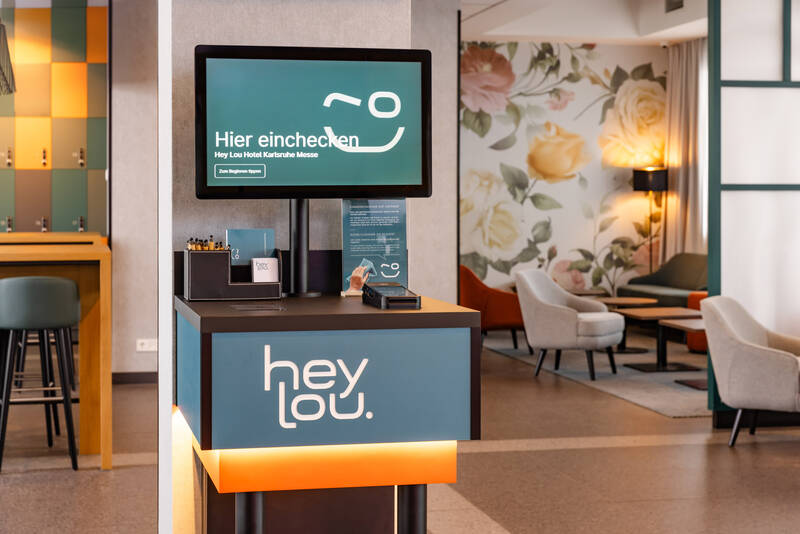 Selbstbedienungs-Check-in-Terminal mit Bildschirm und 'hey lou.' Logo in einem Hotel mit Sitzbereich und Blumenwand im Hintergrund