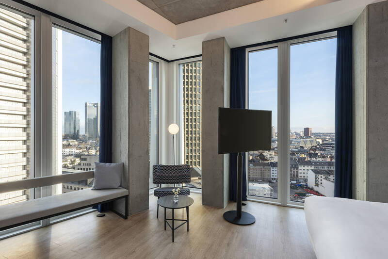 Modern eingerichtetes Zimmer mit großen Fenstern und Blick auf eine Stadt mit Hochhäusern, Sitzbank, Sessel, kleinem Tisch und Fernseher