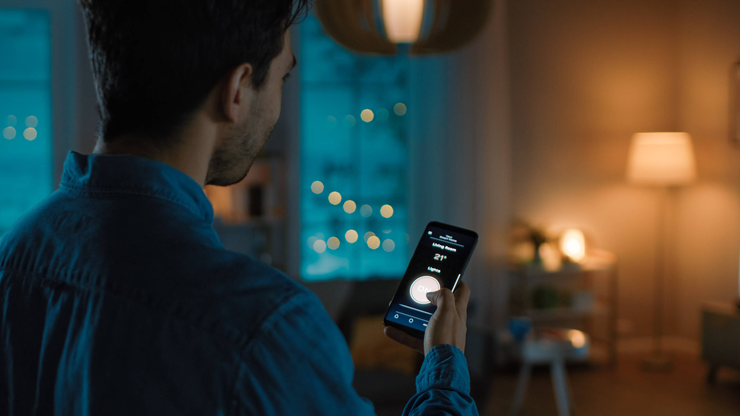Mann hält Smartphone mit Smart-Home-App, zeigt Temperatur und Mondphase, im Wohnzimmer bei Abendlicht