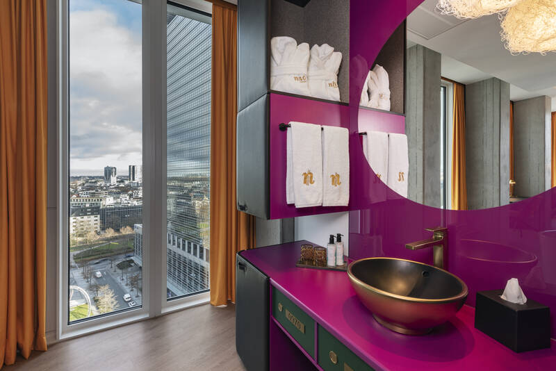 Badezimmer mit großem Fenster und Blick auf Stadt, pinker Waschtisch mit goldfarbenem Waschbecken, runder Spiegel und weißen Handtüchern mit goldenen Initialen