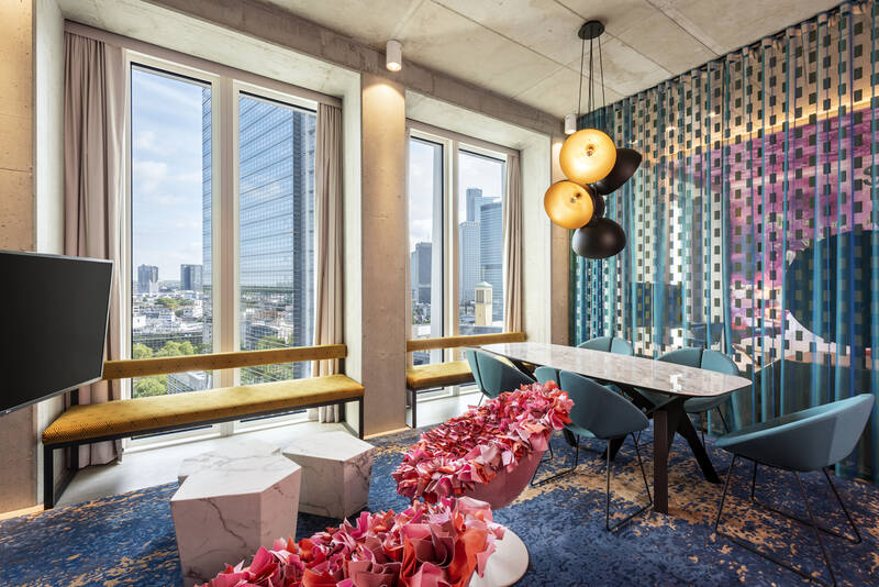 Modernes Hotelzimmer mit großen Fenstern, Blick auf Stadt, Sitzbank mit gelbem Polster, Marmor-Tische, blaue Stühle und bunter Wandgestaltung