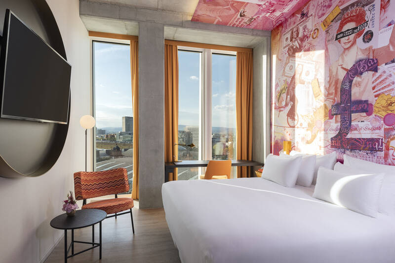Modernes Hotelzimmer mit großem Bett, bunter Wandgestaltung und Blick auf die Stadt durch große Fenster